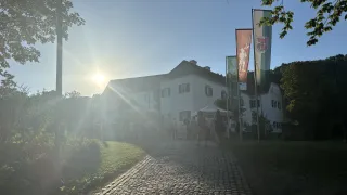 Ein großes weißes Gebäude mit vielen Fenstern steht mit Fahnen an Pfosten davor. Menschen gehen vor ihm bei strahlendem Sonnenschein.