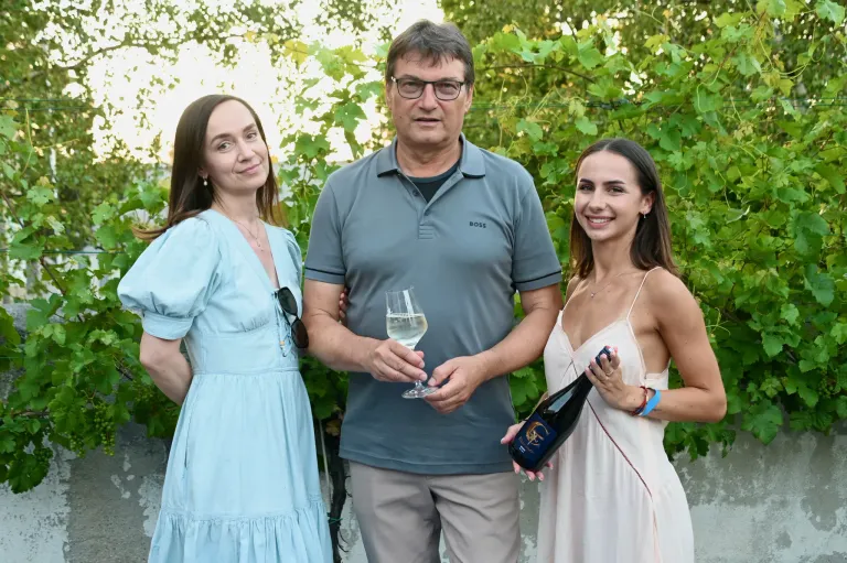Drei Personen stehen vor einem Weinberg. Der Mann hält ein Weinglas und eine Flasche Wein. Die Frauen lächeln.