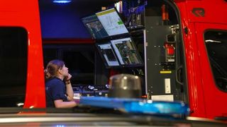 Eine Frau sitzt in einem roten Fahrzeug, wahrscheinlich einem Notfallfahrzeug, mit mehreren Monitoren und einem Bedienfeld vor ihr.