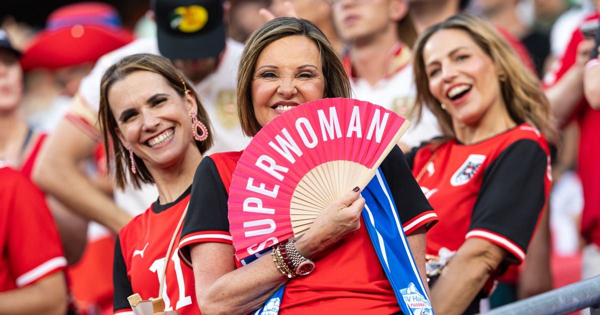 Eine Frau in einem roten Shirt hält einen rosa Fächer mit den Worten 'Super Woman'. Sie lächelt und steht unter anderen Leuten, die ebenfalls lächeln.