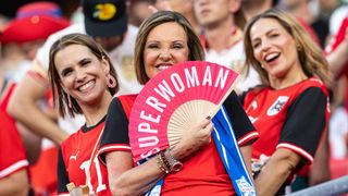 Eine Frau in einem roten Shirt hält einen rosa Fächer mit den Worten 'Super Woman'. Sie lächelt und steht unter anderen Leuten, die ebenfalls lächeln.
