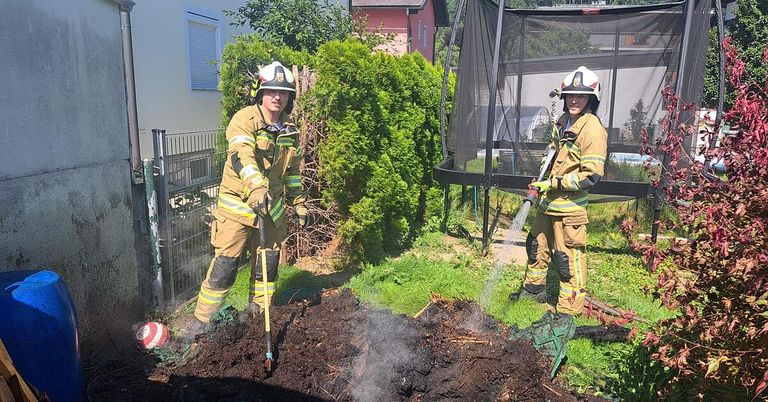 Zwei Feuerwehrleute in voller Ausrüstung spritzen Wasser auf einen rauchigen Grasfleck. Dahinter steht ein Trampolin und ein Haus.