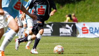Fußballspieler sind auf dem Feld, ein Spieler in einem schwarzen Trikot mit 'Liber' Text kickt einen Fußball. Ein anderer Spieler in einem blauen Trikot läuft auf den Ball zu. Im Hintergrund schauen Menschen zu.
