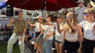 Eine Gruppe von Menschen in passenden weißen Shirts und Röcken steht in einer Reihe unter einem roten Regenschirm. Ein Mann in einem gemusterten Hemd tanzt neben ihnen. Im Hintergrund sitzen Leute auf Treppenstufen.