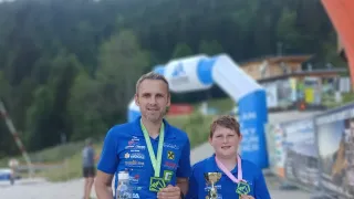 Zwei Personen in blauen Shirts stehen mit ihren Trophäen vor einem unscharfen Hintergrund. Ein Mann hält eine Flasche, und ein Junge lächelt in die Kamera. Dahinter sind ein roter Bogen und Bäume zu sehen.