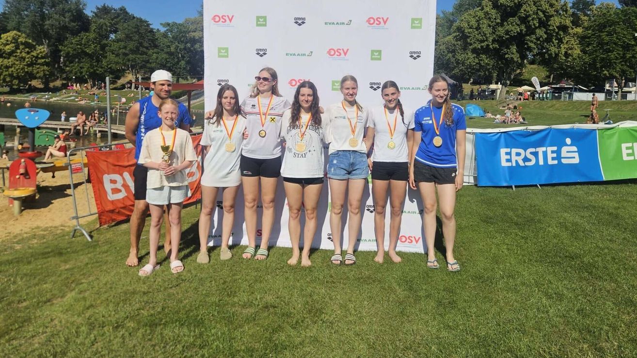 Eine Gruppe junger Frauen und ein Junge stehen zusammen auf einem Grasfeld, lächeln und tragen Medaillen. Sie stehen vor einem weißen Hintergrund mit verschiedenen Logos.