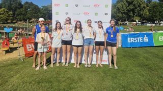 Eine Gruppe junger Frauen und ein Junge stehen zusammen auf einem Grasfeld, lächeln und tragen Medaillen. Sie stehen vor einem weißen Hintergrund mit verschiedenen Logos.