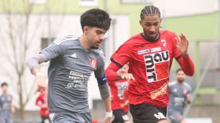 Zwei Männer spielen Fußball, einer in Grau und einer in Rot. Das rote Trikot trägt 'BAU'. Sie rennen in Richtung Ball, andere sind im Hintergrund zu sehen.