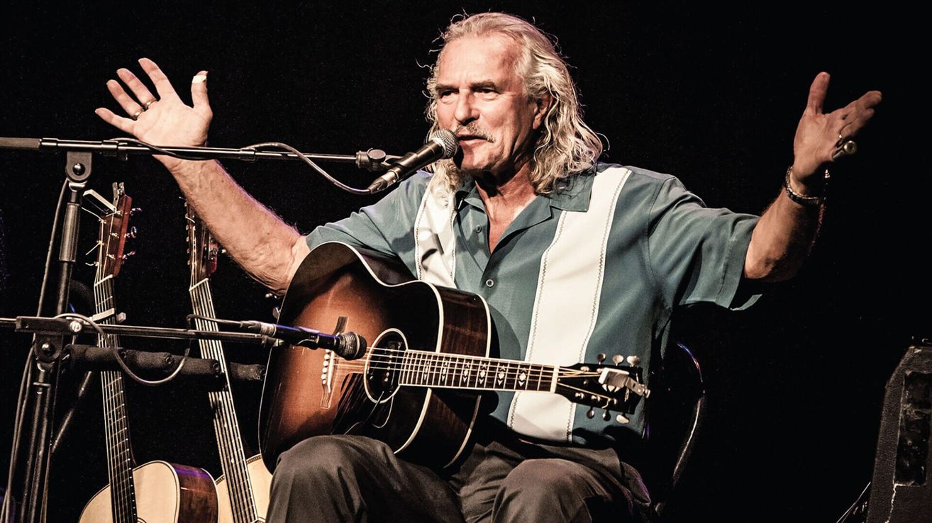 Ein Mann mit langen Haaren und einem Schnurrbart tritt auf der Bühne auf. Er hält eine Gitarre und singt in ein Mikrofon.