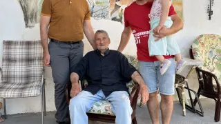 Drei Männer und ein junges Mädchen stehen vor einer Wand mit einem Gemälde. Der Mann in der Mitte sitzt auf einem Stuhl. Der Mann rechts hält ein Kind auf seiner Schulter.