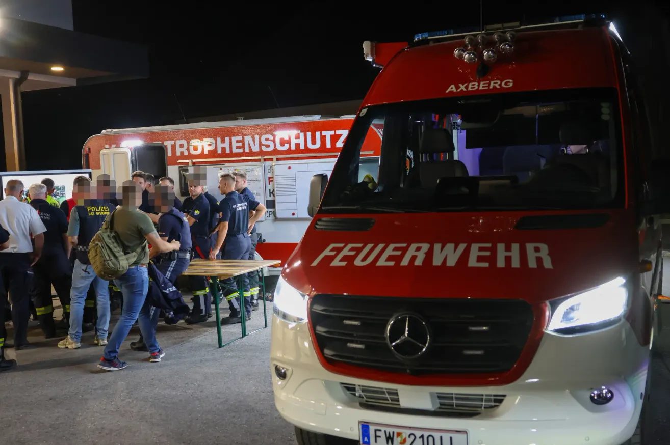 Feuerwehrleute stehen neben einem Feuerwehrauto in der Nacht, einer hält einen Tisch mit verschwommenen Gesichtern. Ein weiteres Fahrzeug, ein rotes Mercedes-Auto, ist davor geparkt.