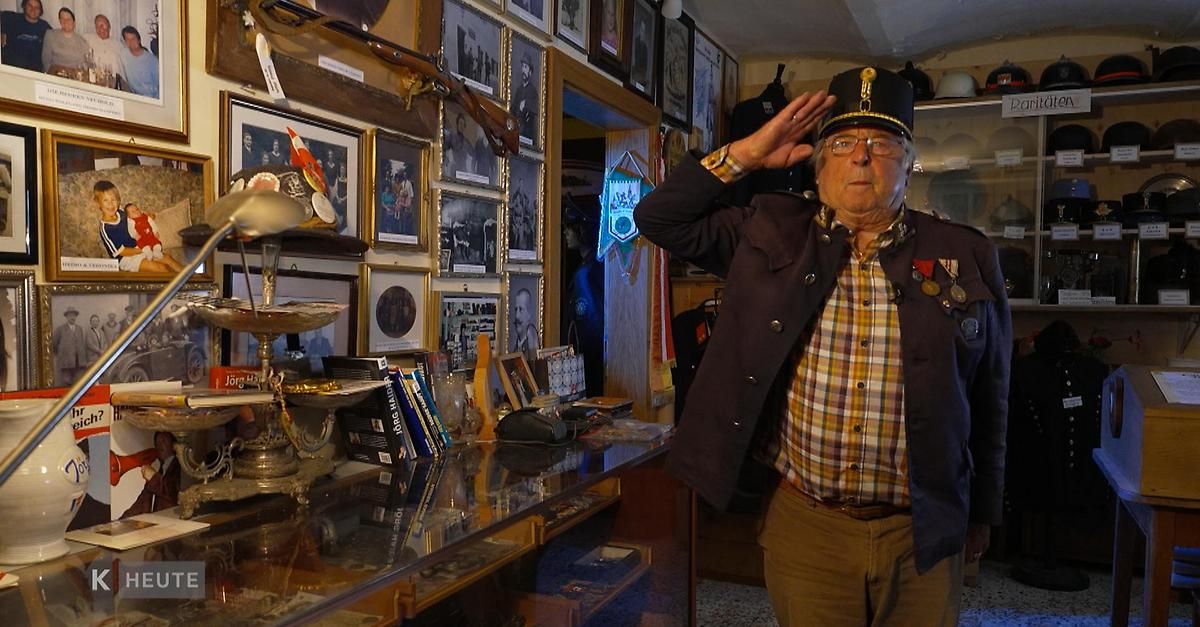 An elderly man salutes in a room adorned with framed photos and military memorabilia. A glass table displays various items.
