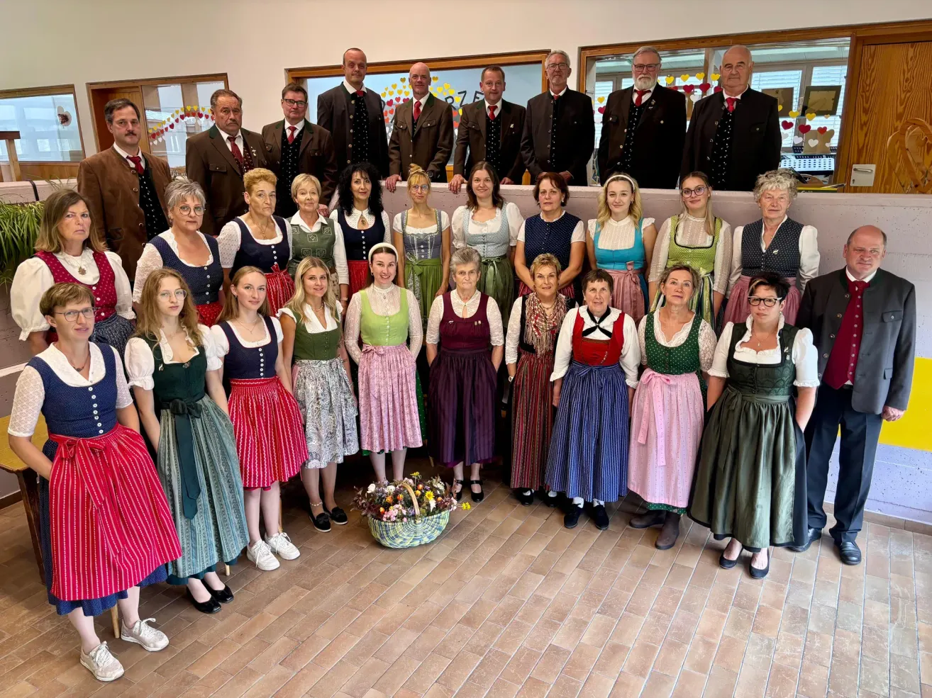 A group of people, both men and women, dressed in traditional clothing, are posing for a photograph. They stand in rows, with some holding a basket of flowers in front.