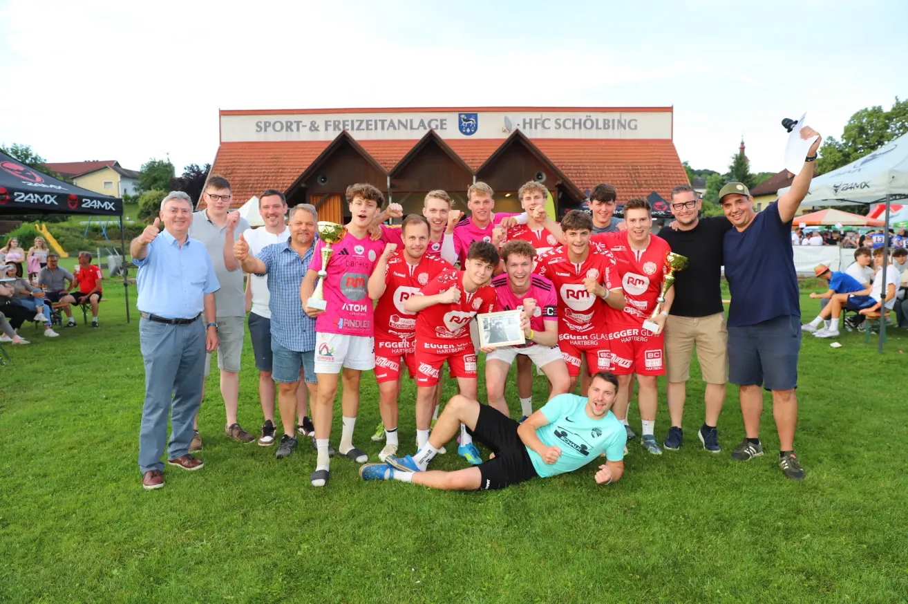 Eine Gruppe von Sportlern, die rote Trikots tragen, posiert mit ihren Trophäen vor einem Gebäude. Ein Mann liegt lächelnd auf dem Gras.
