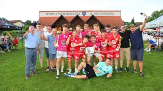 Eine Gruppe von Sportlern, die rote Trikots tragen, posiert mit ihren Trophäen vor einem Gebäude. Ein Mann liegt lächelnd auf dem Gras.