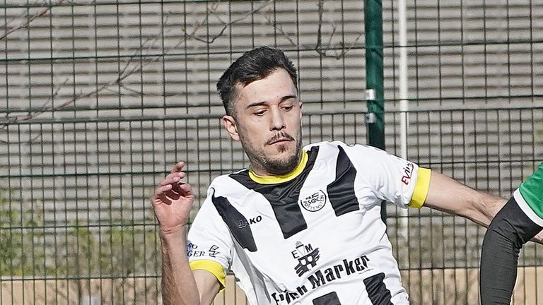 Ein Fußballspieler in einem weißen und schwarzen Trikot steht vor einem Zaun. Er schaut mit konzentriertem Blick nach vorne.