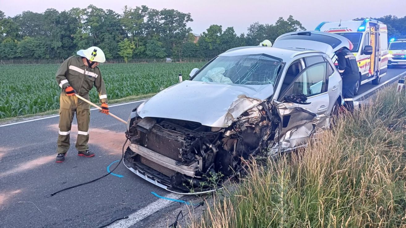 Ein zerbrochenes silbernes Auto steht am Straßenrand, ein Feuerwehrmann hält einen Schlauch in der Nähe der Front.