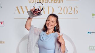 Eine lächelnde Frau hält eine Trophäe in ihrer linken Hand und posiert für ein Foto. Sie ist in einem blauen Kleid und einem silbernen Schal gekleidet. Sie hat kurze Haare und Ohrringe. Der Hintergrund zeigt einen goldenen Text und Zahlen, möglicherweise eine Anerkennungszeremonie.