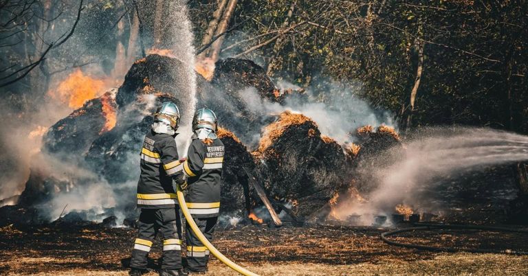 Zwei Feuerwehrleute in Schutzkleidung bekämpfen ein Feuer und spritzen Wasser aus einem Schlauch auf brennendes Material.