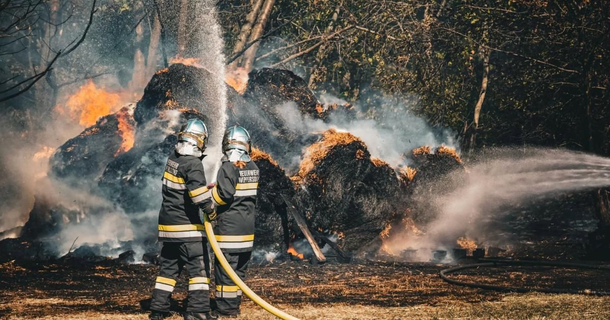 Zwei Feuerwehrleute in Schutzkleidung bekämpfen ein Feuer und spritzen Wasser aus einem Schlauch auf brennendes Material.