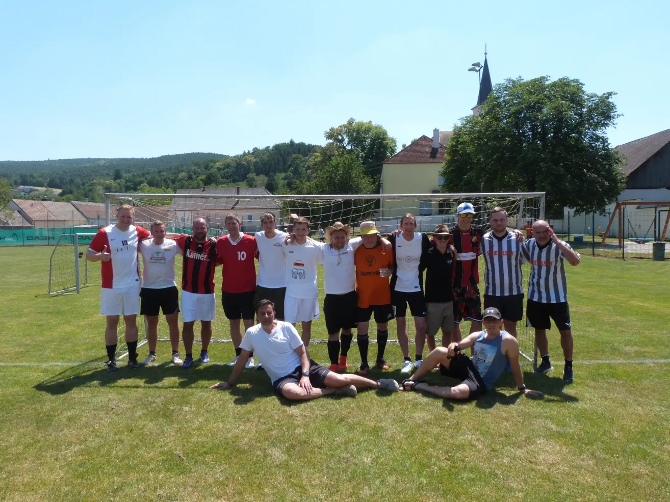 Eine Gruppe von Männern in Sportausrüstung, stehend und sitzend vor einem Fußballtor auf einem Rasenfeld mit Häusern und Bäumen im Hintergrund.
