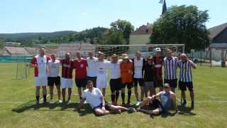 Eine Gruppe von Männern in Sportausrüstung, stehend und sitzend vor einem Fußballtor auf einem Rasenfeld mit Häusern und Bäumen im Hintergrund.