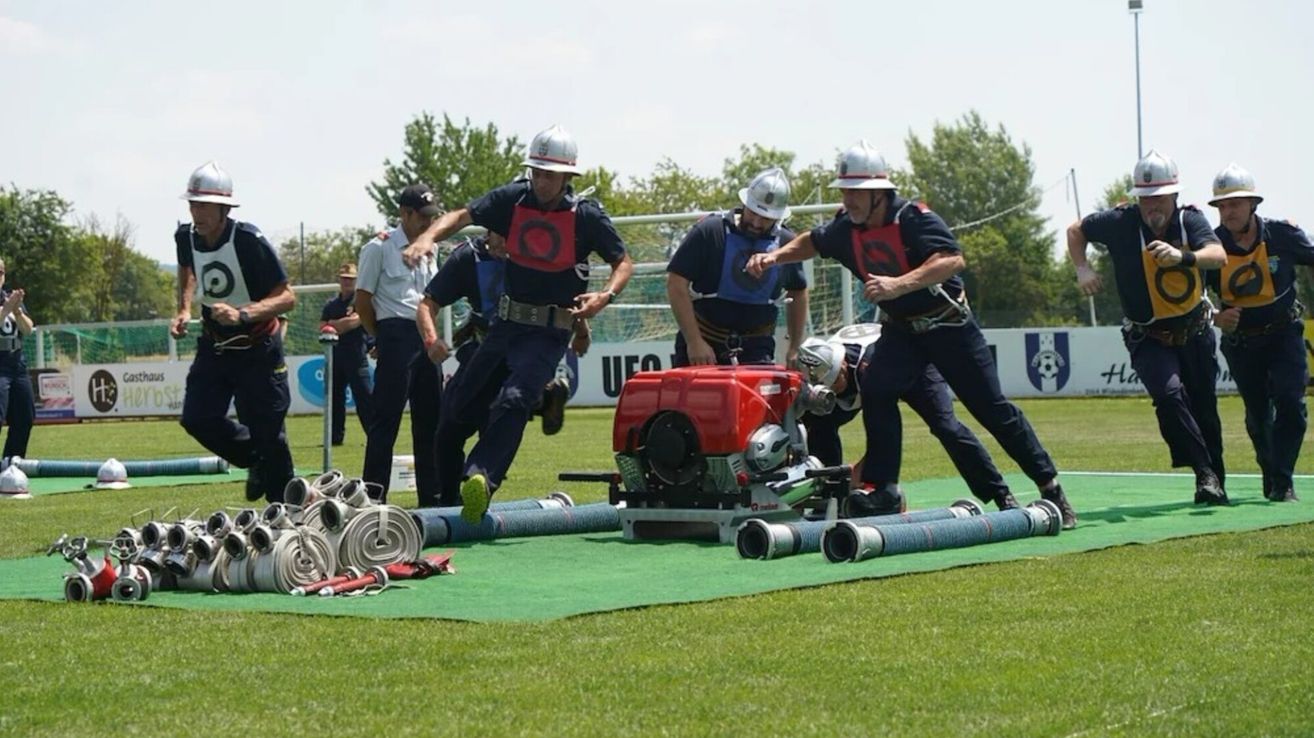 Feuerwehrleute rennen und springen über Hindernisse auf einem Feld, tragen Helme und Uniformen. Eine rote Maschine steht auf dem Boden.
