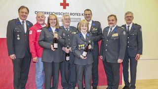 Eine Gruppe von Menschen in Militäruniformen, die Flaschen Wein halten, posiert für ein Foto vor einem rot-weißen Hintergrund.