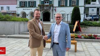 Zwei Männer schütteln sich die Hände vor einem Gebäude mit dem Wort Rathaus. Sie tragen Anzüge, einer in Blau und der andere in Braun. Hinter ihnen sind eine Holzbank, ein Baum und eine Treppe zu sehen.