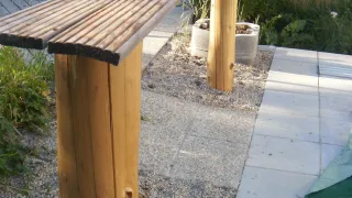 Ein Außenbereich mit Holzbänken und -pfosten. Ein Metallständer und ein blauer Tisch sind in der Nähe. Ein Betonpflanzer beherbergt Grünpflanzen.