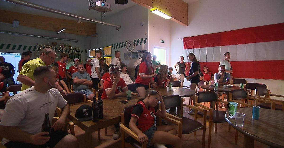 Ein Raum mit Menschen, die um Tische herum sitzen und ein Fußballspiel auf einer Projektionsleinwand verfolgen. Einige tragen rote Trikots. Flaschen und Gläser stehen auf den Tischen.