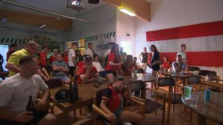 Ein Raum mit Menschen, die um Tische herum sitzen und ein Fußballspiel auf einer Projektionsleinwand verfolgen. Einige tragen rote Trikots. Flaschen und Gläser stehen auf den Tischen.