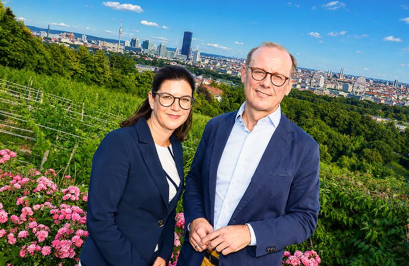 Ein Mann und eine Frau in Geschäftskleidung stehen in einem Blumenfeld und blicken auf eine Stadt.