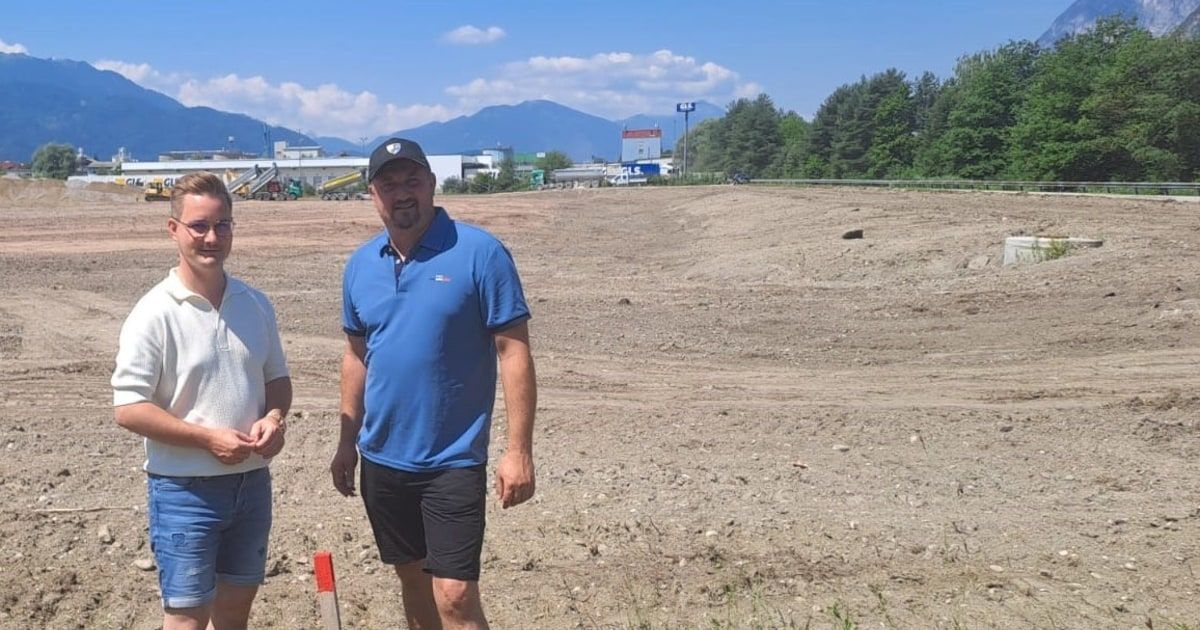 Zwei Männer stehen auf einem schlammigen Feld, einer in einem blauen Poloshirt und der andere in einem weißen Shirt. Hinter ihnen befindet sich eine bergige Landschaft und ein großes Gebäude mit einem roten Dach.