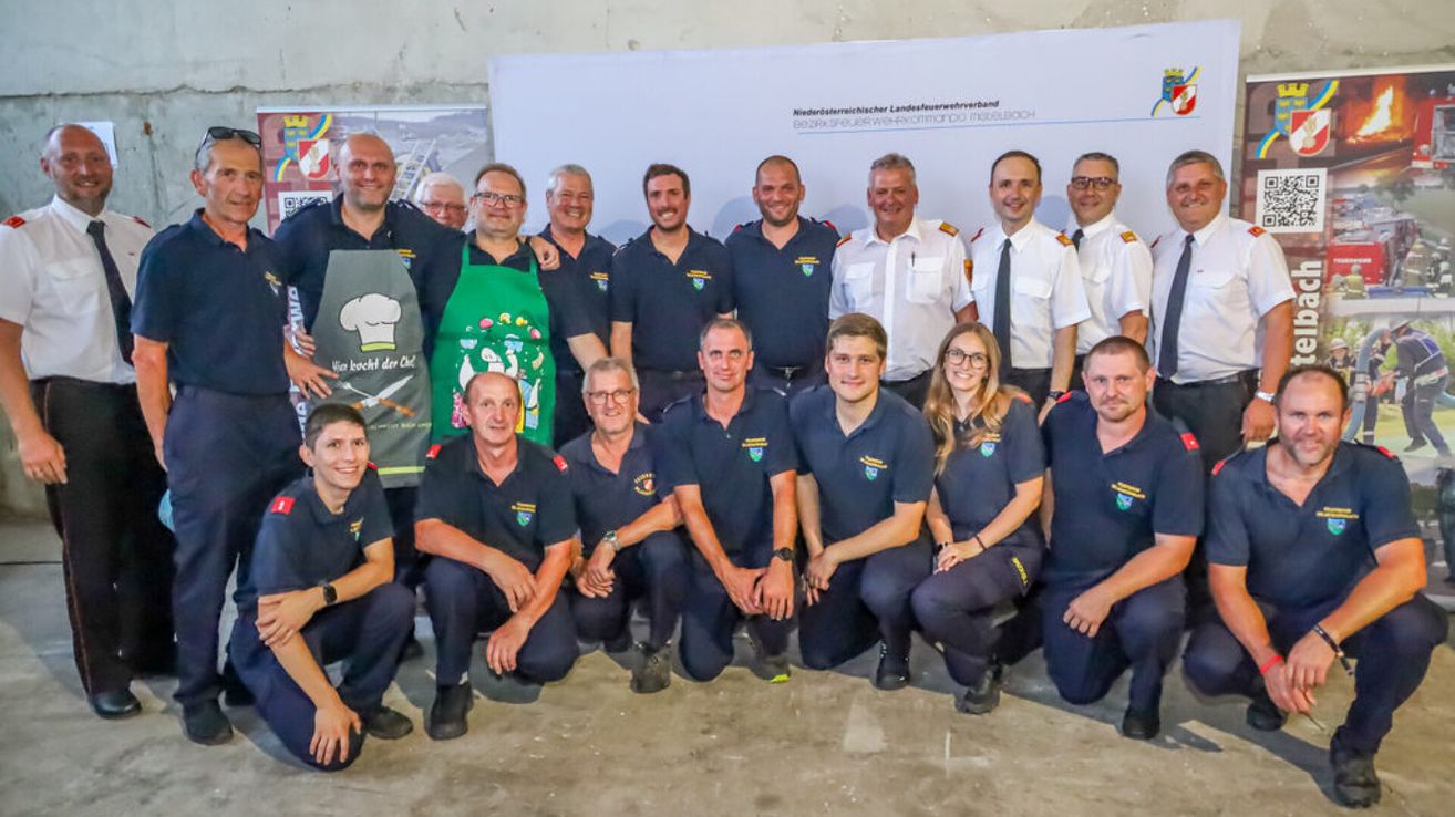 Eine Gruppe von Feuerwehrleuten posiert vor einem Banner. Zwei tragen Schürzen, einer hat eine Kochmütze auf. Sie lächeln.