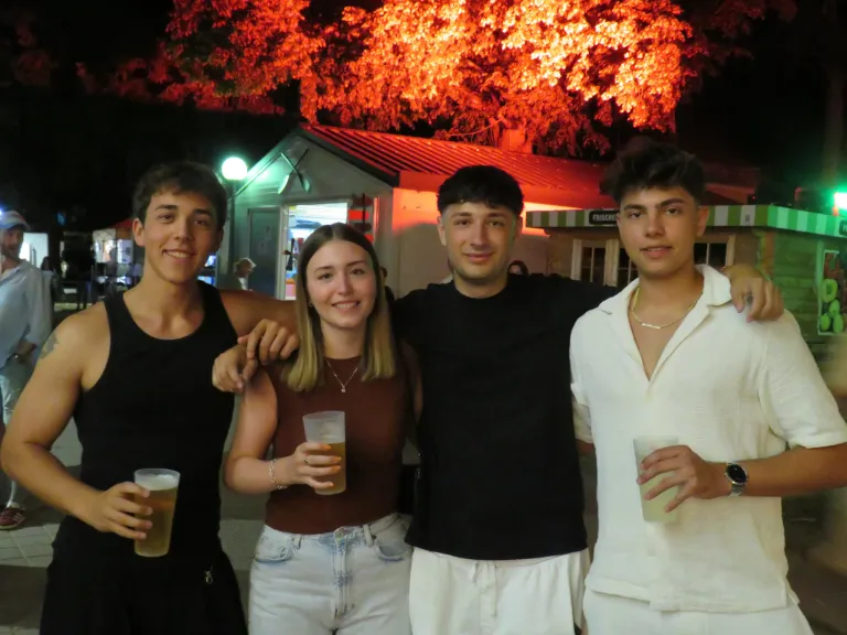 Vier Freunde posieren für ein Foto draußen bei Nacht, mit einem leuchtenden Baum und einem Gebäude dahinter. Jeder hält ein Glas mit Getränk.