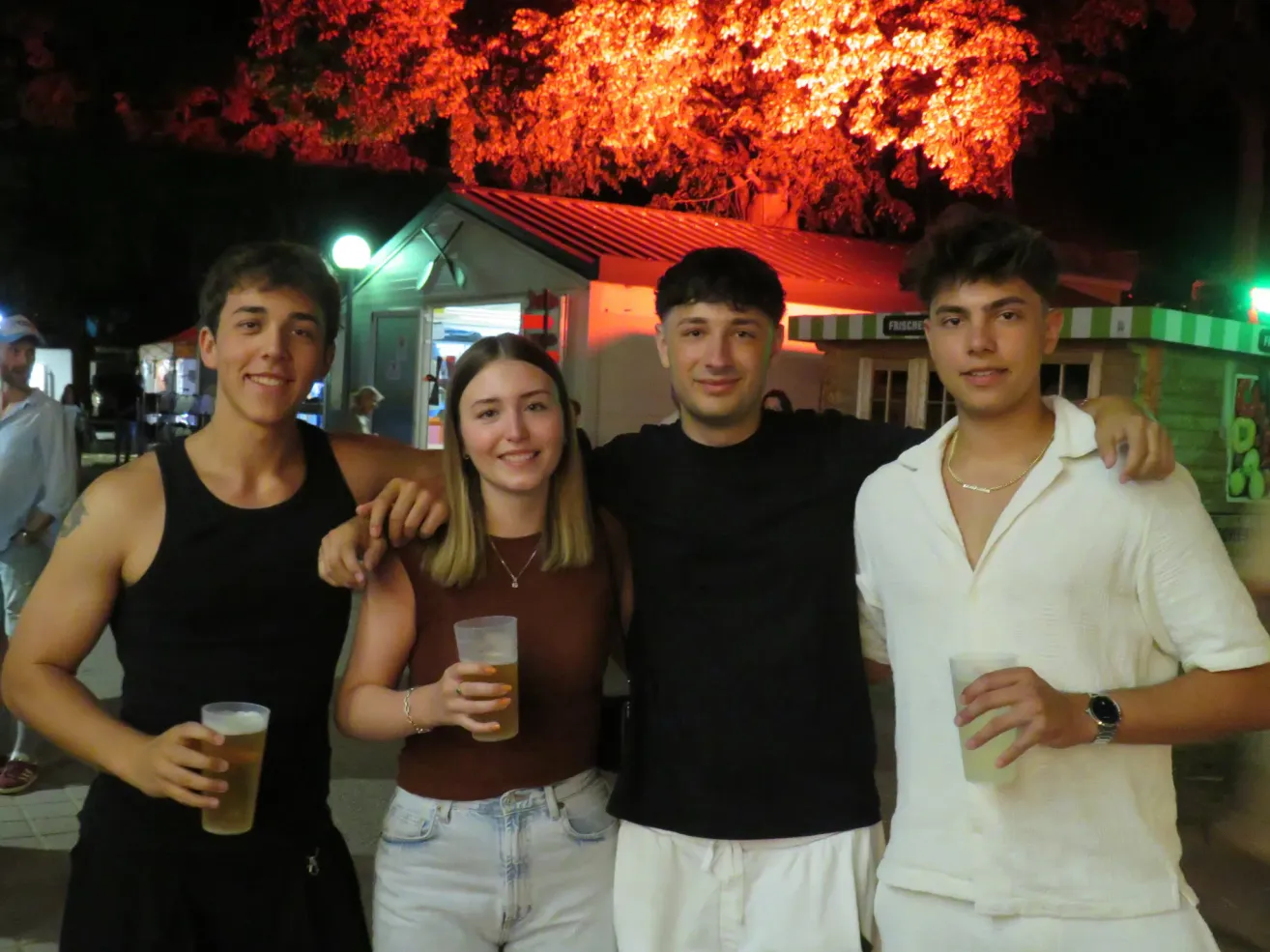 Vier Freunde posieren für ein Foto draußen bei Nacht, mit einem leuchtenden Baum und einem Gebäude dahinter. Jeder hält ein Glas mit Getränk.