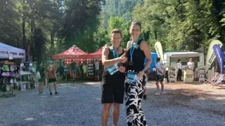 Zwei Frauen stehen auf einem Podium mit einem blauen Schild bei einer Outdoor-Veranstaltung, tragen Medaillen und lächeln. Dahinter sind verschiedene Zelte und Menschen verstreut.