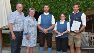 Fünf Personen stehen zusammen und sind in traditioneller Kleidung gekleidet. Zwei Frauen tragen gemusterte Kleider, und der Mann in der Mitte trägt eine blaue Weste mit gelber Krawatte. Hinter ihnen befindet sich eine Holzbank und ein grüner Hintergrund.