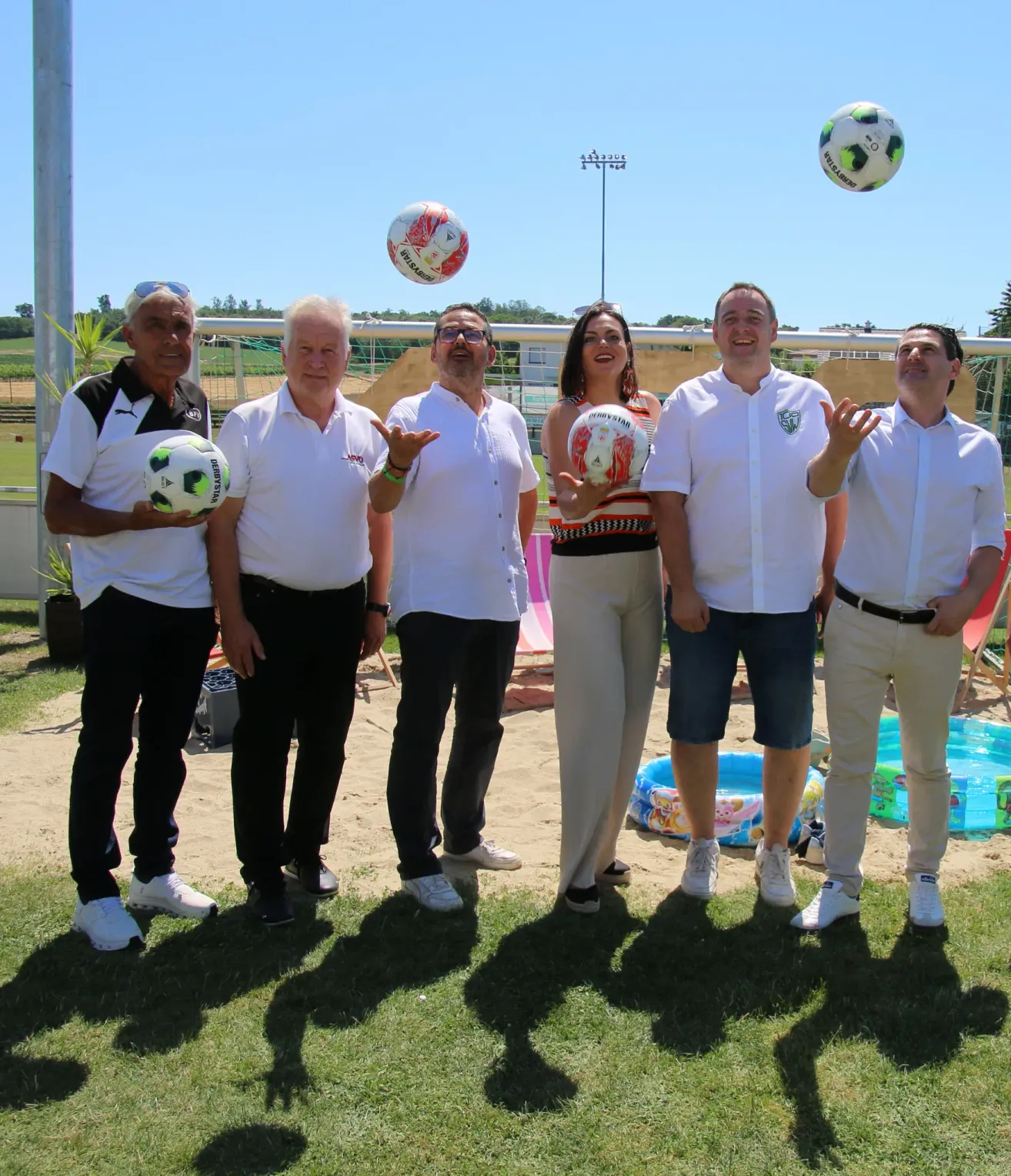 A group of people in white shirts and pants are standing on the grass. Two soccer balls are in the air and one is held by a person.