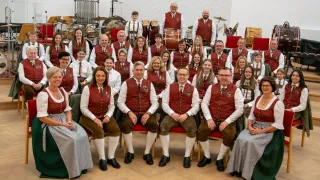 Eine Gruppe von Musikern in roten Westen und traditionellen Kostümen, sitzend und stehend, mit verschiedenen Musikinstrumenten im Hintergrund.