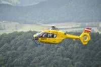 A yellow helicopter flies over a dense forest with mountains in the background, displaying a red and white flag.