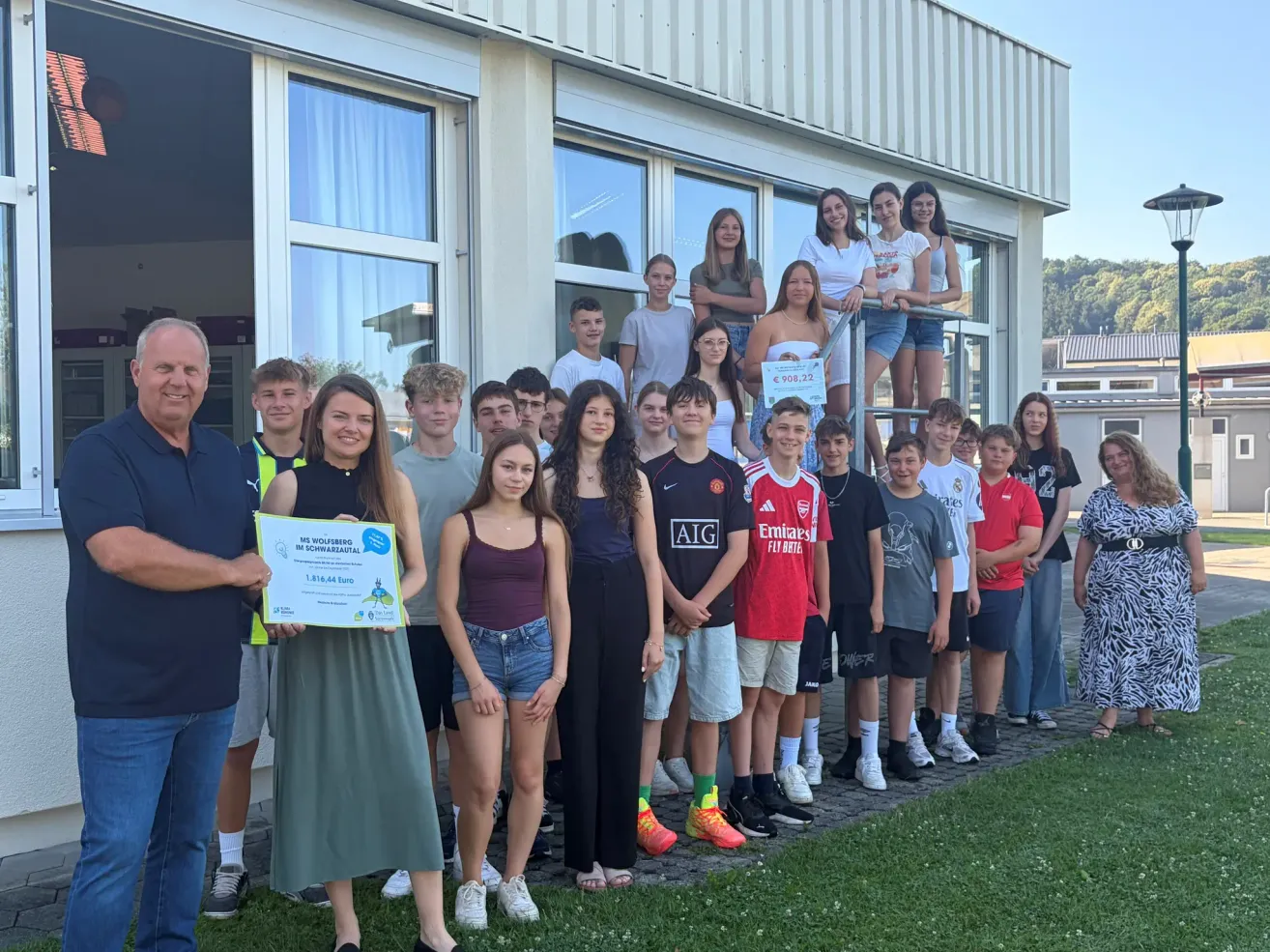 Eine Gruppe von Schülern und Mitarbeitern posiert für ein Foto vor einem Gebäude und hält ein Zertifikat. Das Zertifikat wird von einer Frau in einem grünen Kleid präsentiert.