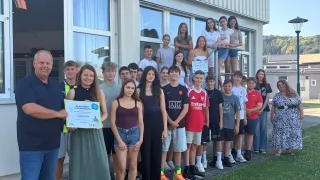 Eine Gruppe von Schülern und Mitarbeitern posiert für ein Foto vor einem Gebäude und hält ein Zertifikat. Das Zertifikat wird von einer Frau in einem grünen Kleid präsentiert.
