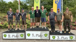 Eine Gruppe von Männern steht auf einem Podest mit Medaillen um den Hals. Das Podest hat Schilder für 1., 2. und 3. Platz. Sie lächeln und posieren für ein Foto.