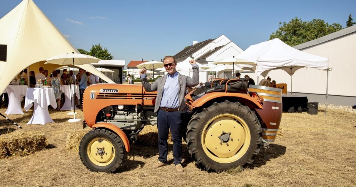 Ein Mann in einer Sonnenbrille und einem Anzug posiert neben einem orangefarbenen Traktor, mit Zelten und Häusern im Hintergrund.