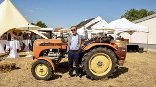Ein Mann in einer Sonnenbrille und einem Anzug posiert neben einem orangefarbenen Traktor, mit Zelten und Häusern im Hintergrund.