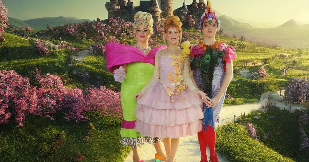 Drei Frauen in farbenfrohen Kostümen stehen in einem Garten, eine in einem pinkfarbenen Kleid mit Blumen, eine andere in Grün mit einer rosa Schleife und die dritte in einem gefiederten Outfit. Hinter ihnen bilden ein Schloss und rosa Blumen eine märchenhafte Szene.