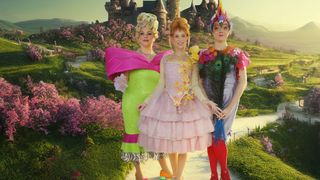 Drei Frauen in farbenfrohen Kostümen stehen in einem Garten, eine in einem pinkfarbenen Kleid mit Blumen, eine andere in Grün mit einer rosa Schleife und die dritte in einem gefiederten Outfit. Hinter ihnen bilden ein Schloss und rosa Blumen eine märchenhafte Szene.