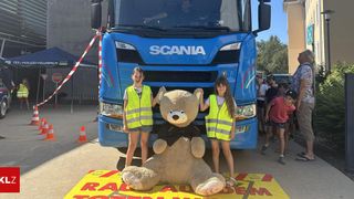 Zwei Mädchen in Warnwesten stehen neben einem großen Teddybären vor einem Scania-Lkw. Eine Menschenmenge von Kindern beobachtet von hinten.